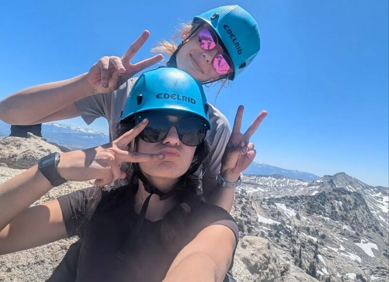Two women wearing climbing helmets and sunglasses pose for a photo on a rocky mountaintop. They are both making peace signs with their hands. The sky is blue and clear, and there are mountains in the background. The women appear to be enjoying their time outdoors.